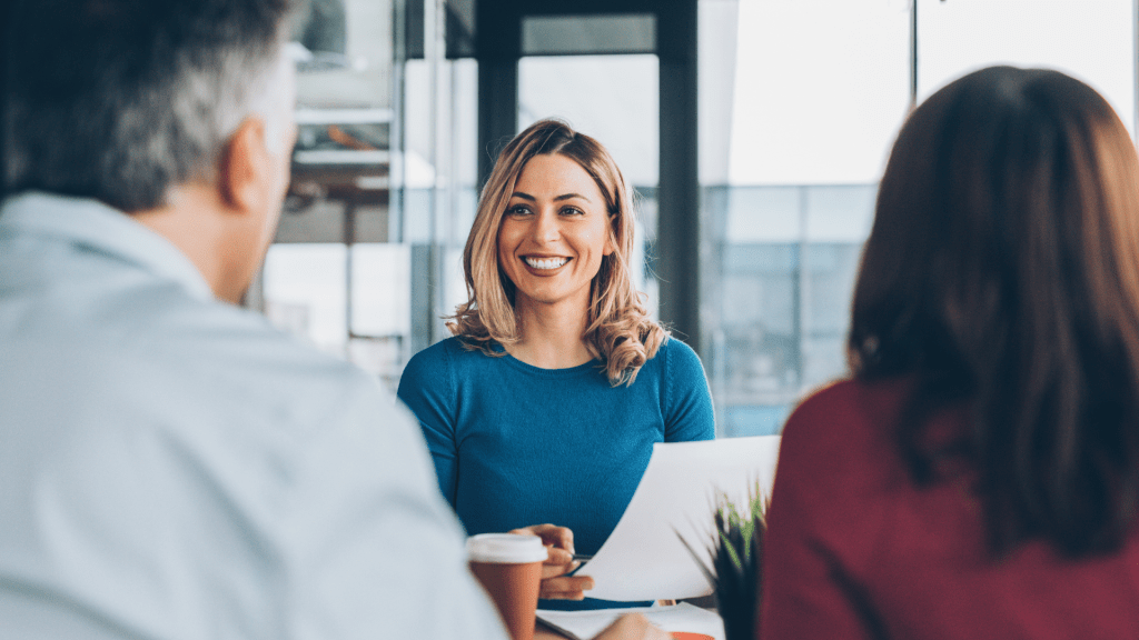 An insurance agent smiles warmly while meeting with two clients across a table in a bright, modern office setting