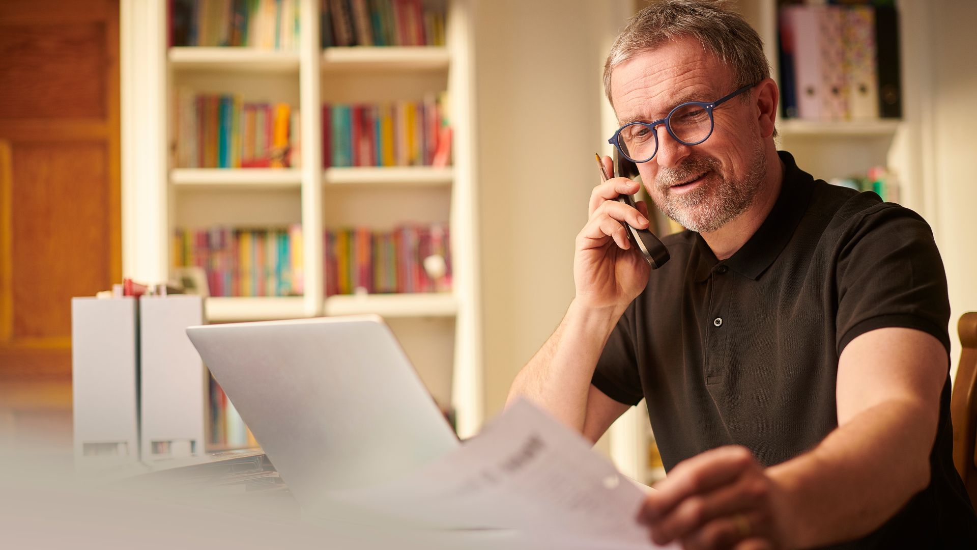 A small business owner talks on the phone while reviewing paperwork at a home office desk with a laptop open in front of him